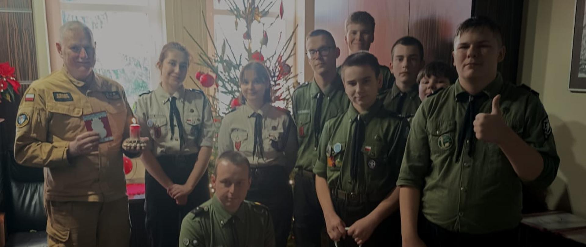 Grupowe zdjęcie harcerzy i funkcjonariusza Państwowej Straży Pożarnej z Betlejemskim Światłem Pokoju; uczestnicy stoją przed choinką w pomieszczeniu służbowym.