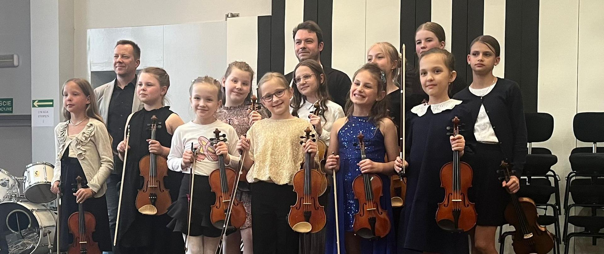 Dwóch mężczyzn i jedenaścioro dziewczynek stoją na scenie ze skrzypcami.
