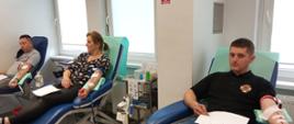 Na zdjęciu dwaj mężczyzna po lewej w szarej koszulce i czarnych spodniach w środku kobieta w bluzce czarnej w kolorowe kwiaty i w czarnych spodniach po prawej mężczyzna w czarnej koszulce i dżinsach siedzą na granatowych fotelach oddają krew w pomieszczeniu o białych ścianach i szarej posadzce w Regionalnym Centrum Krwiodawstwa i Krwiolecznictwa w Oddziale Terenowym w Gorlicach.