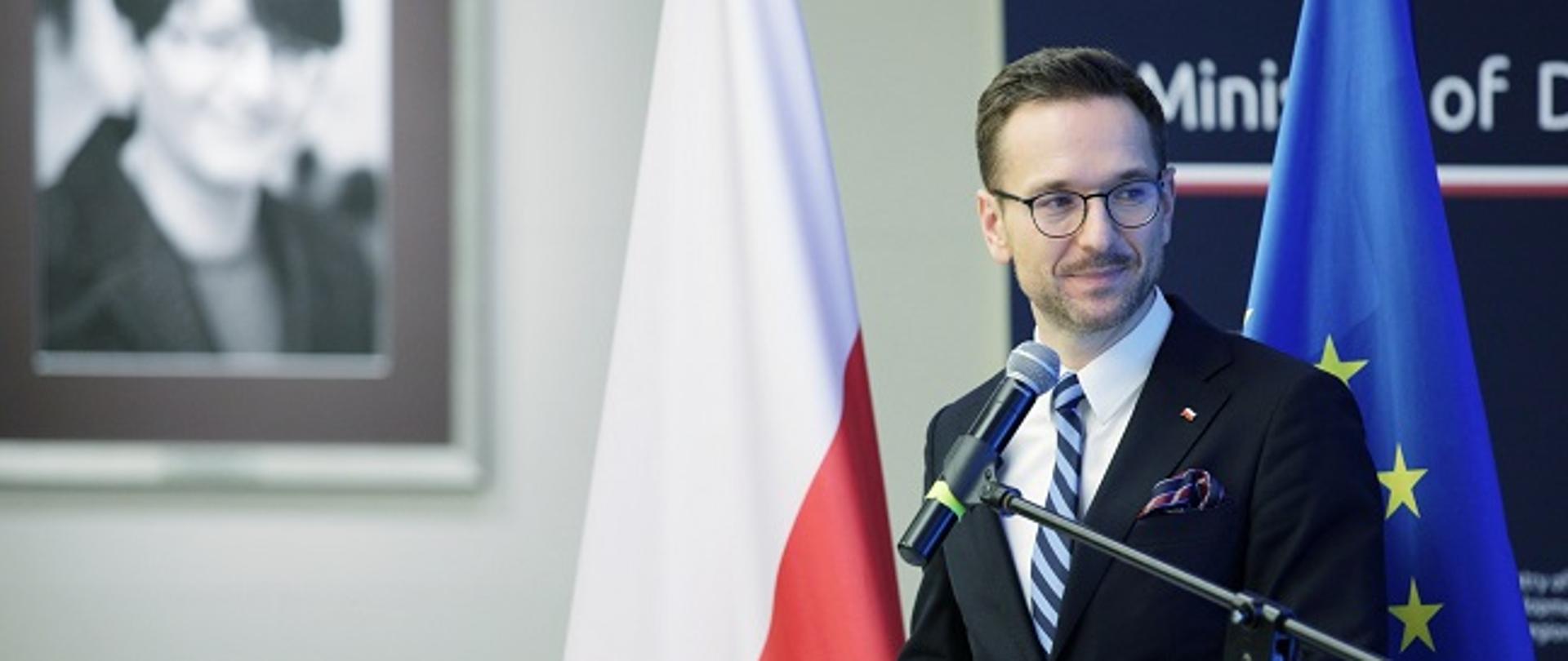 Wiceminister Waldemar Buda stoi przy mównicy z mikrofonem. W tle flagi Polski i UE