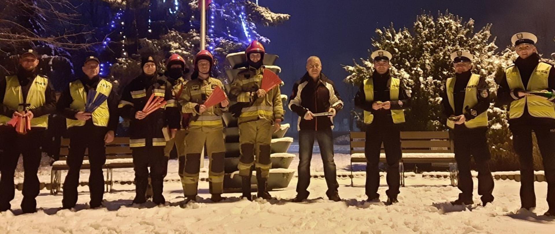 Na zdjęciu na tle ubranej choinki stoją przedstawiciele służb mundurowych trzymający elementy odblaskowe przeznaczone dla mieszkańców powiatu myszkowskiego