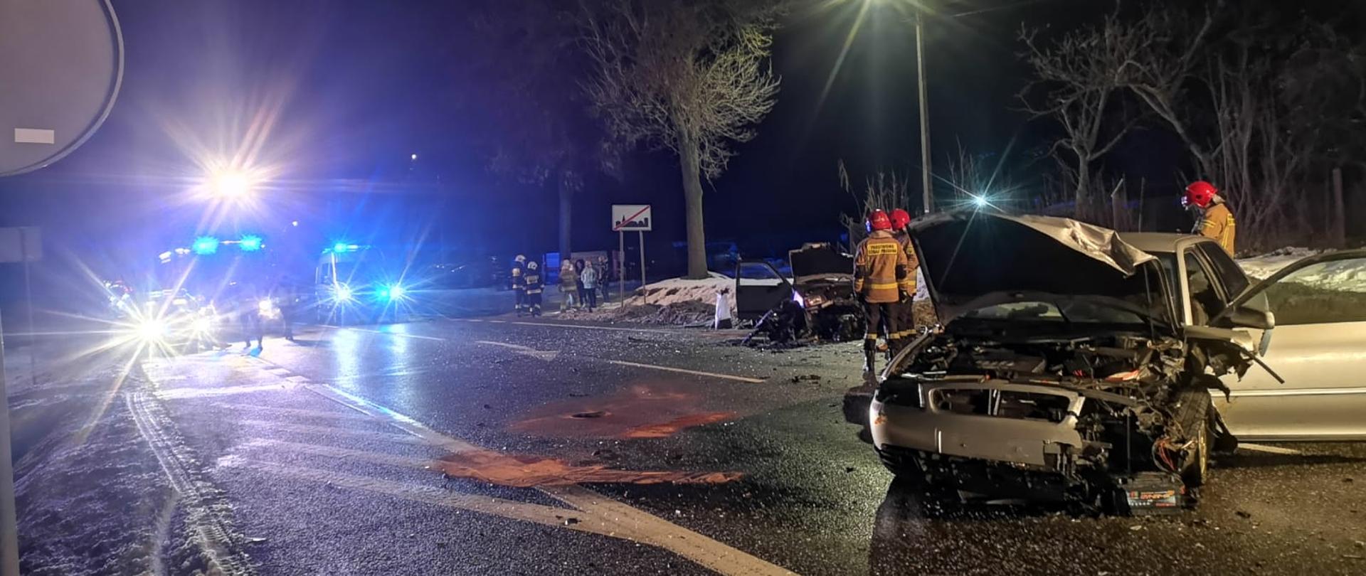 Zdjęcie przedstawia na której, po prawej stronie stoją dwa zniszczone auta. Po lewej, w pewnym oddaleniu stoją samochody służb ratowniczych. Przy zniszczonych autach stoi trzech strażaków.