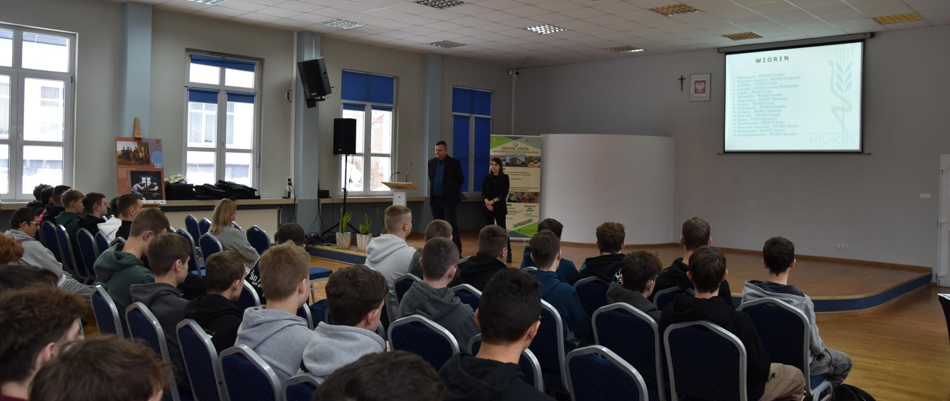 Grupa uczniów siedząca w dużej jasnej sali. Przed nimi stoi kobieta i mężczyzna za którymi wyświetlana jest prezentacja. Po lewej stronie w pomieszczeniu znajdują się okna
