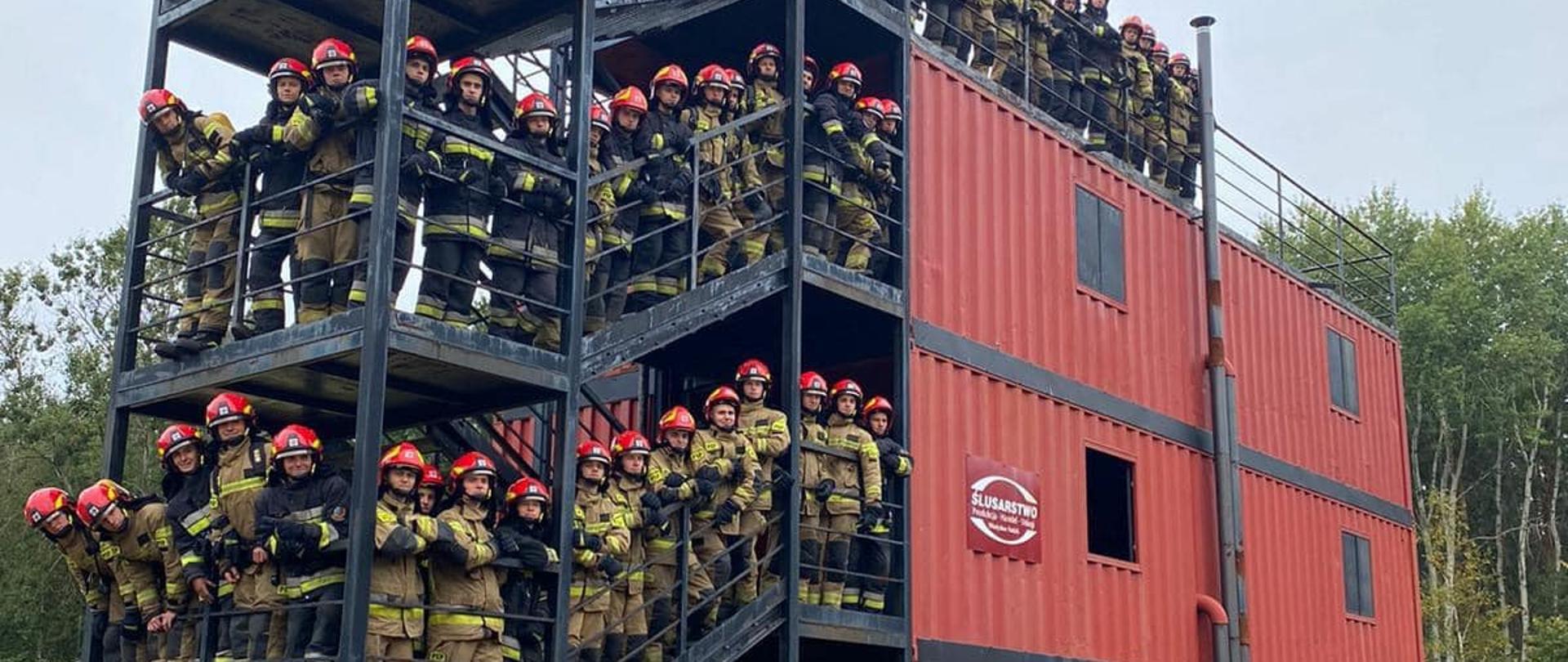 Komora rozgorzeniowa na schodach której ustawieni są strażacy biorący udział w honorowej wspinaczce