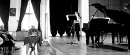 czarno białą fotografia po prawej stronie siwy starszy i muzycy na scenie przy czarnym fortepianie człowiek po lewej grupa osób słuchająca wykładu