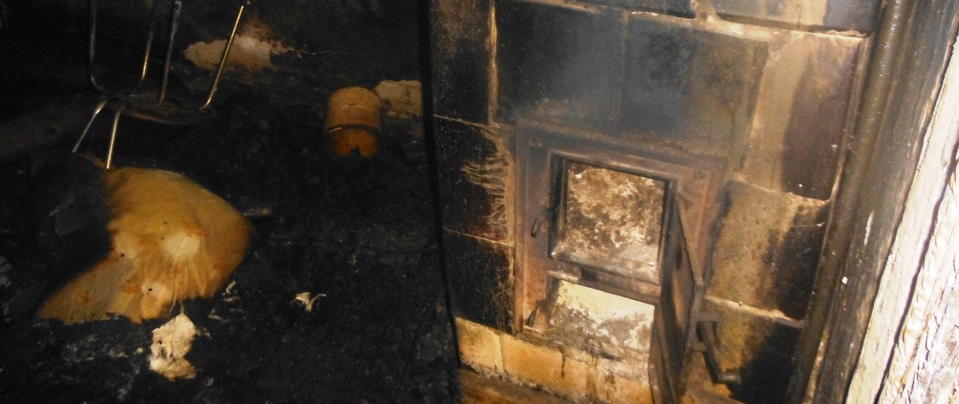 Zadymione wnętrze pokoju. Okopcony piec kaflowy z otwartymi drzwiczkami.. Na ziemi porozrzucane nadpalone przedmioty – krzesło, poduszka, garnek.