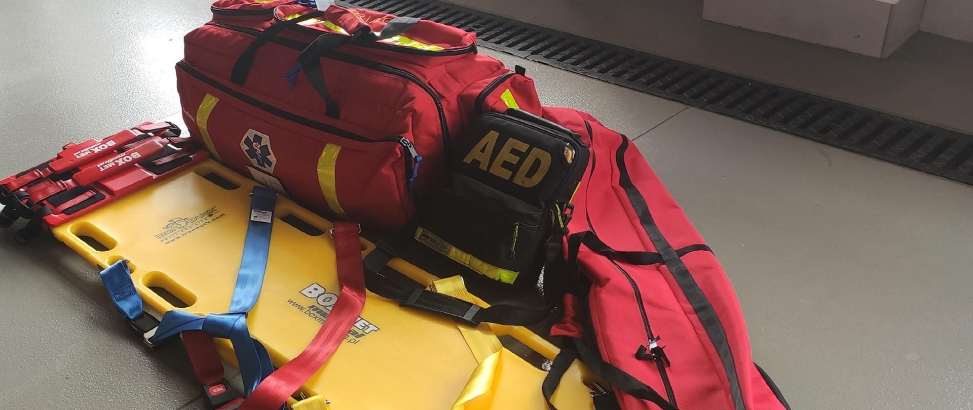 Zdjęcie obrazuje nosze typu deska ortopedyczna walizkę medyczną, szyny do stabilizacji,AED