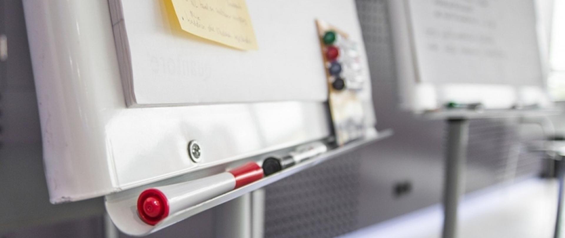 Dwie białe tablice z markerami i żółtą karteczką w pomieszczeniu edukacyjnym lub biurowym.