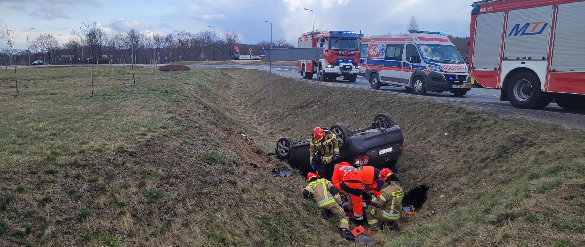W rowie, przy drodze asfaltowej na dachu leży samochód osobowy, w kolorze grafitowym. Przy samochodzie znajduje się 3 strażaków i dwóch ratowników medycznych udzielających pierwszej pomocy osobie poszkodowanej. Na drodze asfaltowej soją dwa samochody strażackie, a między nimi znajduje się karetka pogotowia.