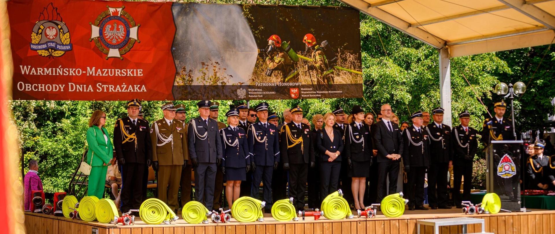 Duża scena pod zadaszeniem. Na scenie strażacy, funkcjonariusze, żołnierze w mundurach galowych oraz zaproszeni goście. Słonecznie.