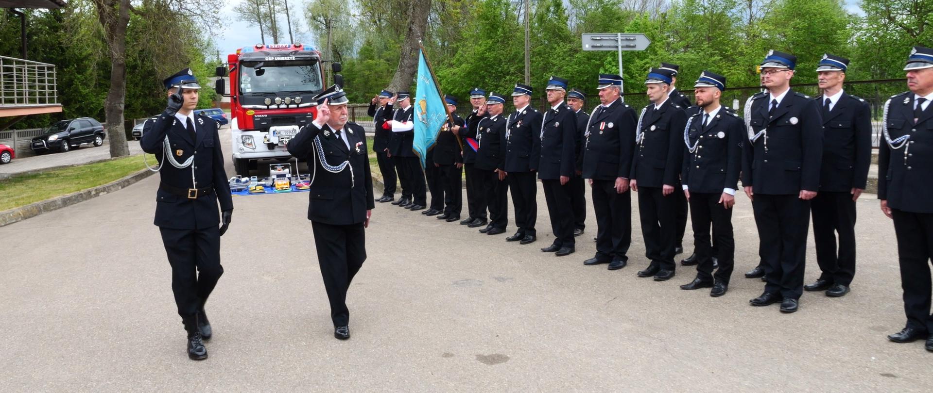 Strażacy w galowych mundurach maszerują podczas uroczystego apelu na placu przed remizą, w tle widoczny czerwony wóz strażacki i poczet sztandarowy.