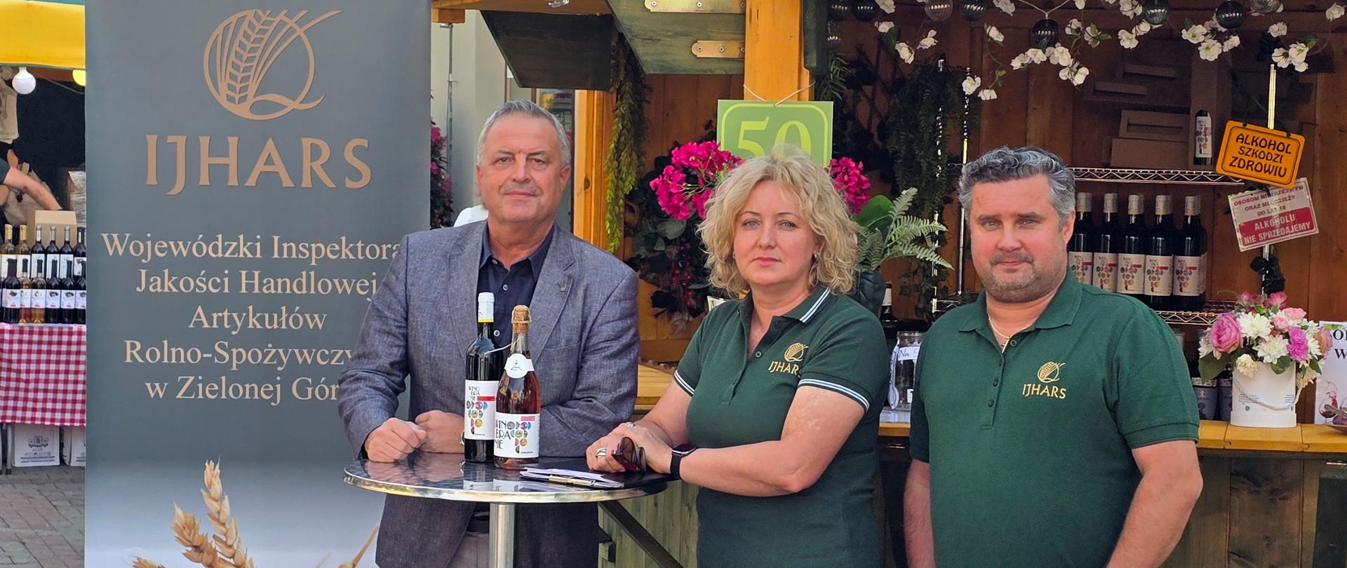 Zdjęcie przedstawia Inspektorów Wojewódzkiego Inspektoratu Jakości handlowej Artykułów Rolno-Spożywczych w Zielonej Górze w towarzystwie Zastępcy Wojewódzkiego Inspektora ubranych w zielone koszulki polo z logiem WIJHARS. Za inspektorami mozemy dostrzec budkę winiarską, na stoliku 2 wina z etykietą Winobranie 2025 oraz roll-up z logiem IJHARS i napisem Wojewódzki Inspektorat Jakości handlowej Artykułów Rolno-Spożywczych w Zielonej Górze.