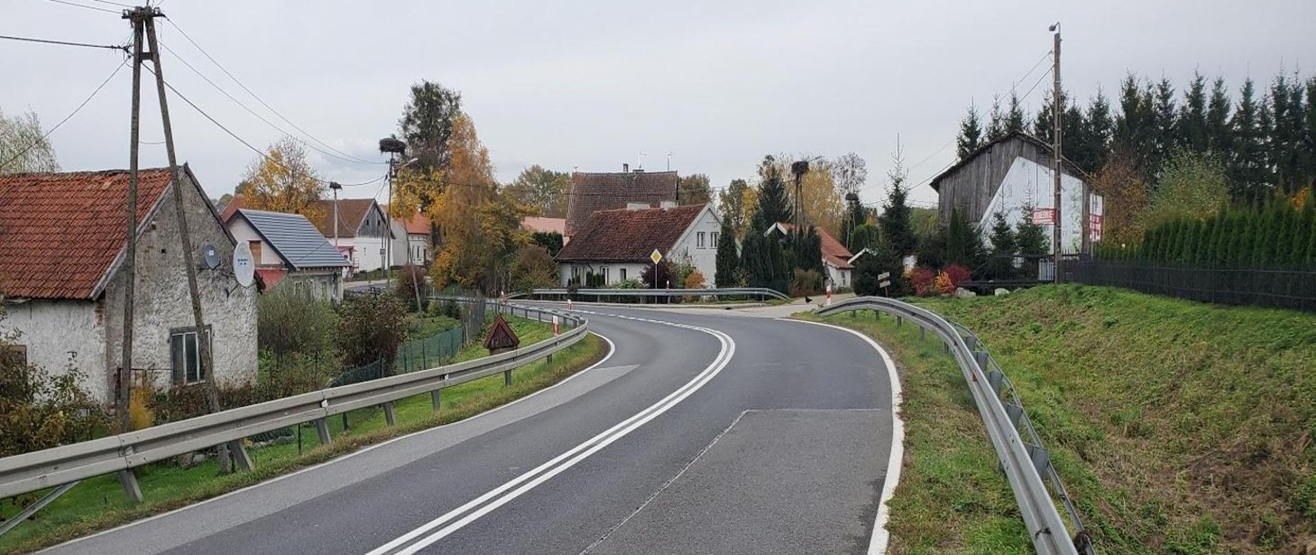 Zakręt drogi, przebiegającej przez małą miejscowość. Widoczne liczne zabudowania.