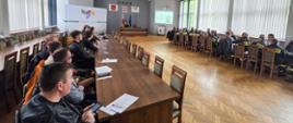 Zdjęcie przedstawia siedzących przy stołach strażaków z jednostek OSP w ubraniach koszarowych. Na samym końcu i środku siedzi przewodniczący komisji przy laptopie. Nad nim wisi godło Polski i miasta Ustrzyki Dolne, po lewej na dużym banerze widać logo Ustrzyk Dolnych a po prawej stronie widać wyświetlane slajdy z rzutnika. Strażacy OSP siedzą w dwóch rzędach biurek, połowa po prawej a druga połowa po lewej stronie sali. Mają Oni przed sobą położone na stołach kartki i długopisy. 