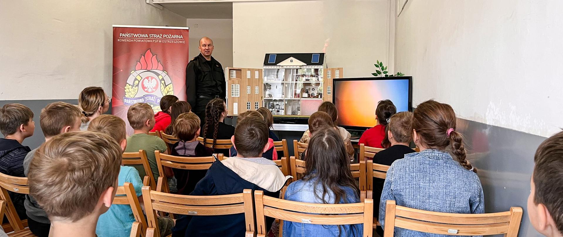 Zdjęcie w trakcie pokazu mobilnego symulatora zagrożeń pożarowych w postaci budynku jednorodzinnego. Uczniowie siedzą na krzesłach tyłem do fotografa. Na dalszym planie prelegent omawia przebieg symulacji.