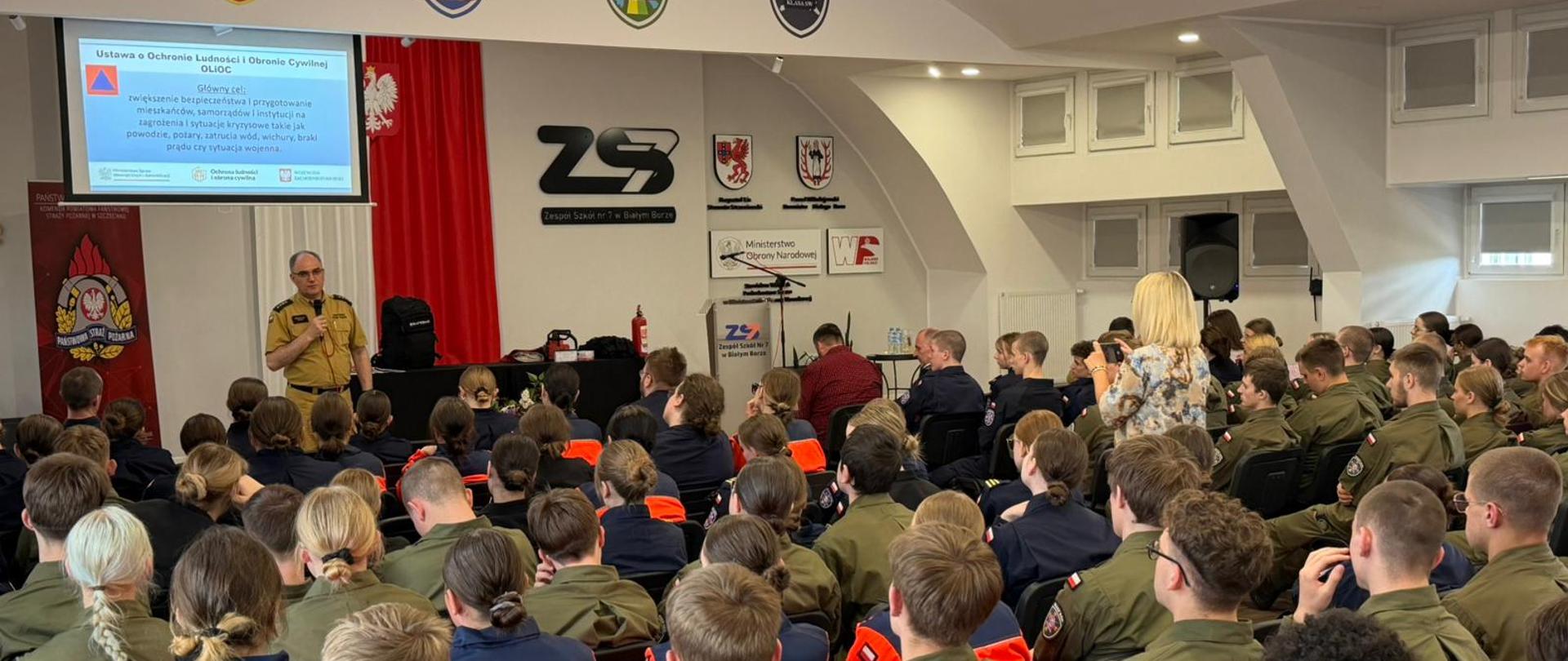 W Zespole Szkół nr 7 w Białym Borze odbywa się spotkanie komendanta powiatowego PSP w Szczecinku, st. kpt. Mirosława Śledzia, z uczniami klas mundurowych. Funkcjonariusz w umundurowaniu Państwowej Straży Pożarnej przemawia do siedzących na sali uczniów. W tle widoczny jest ekran z prezentacją dotyczącą ustawy o ochronie ludności i obronie cywilnej, baner PSP oraz biało-czerwone dekoracje z godłem Polski.