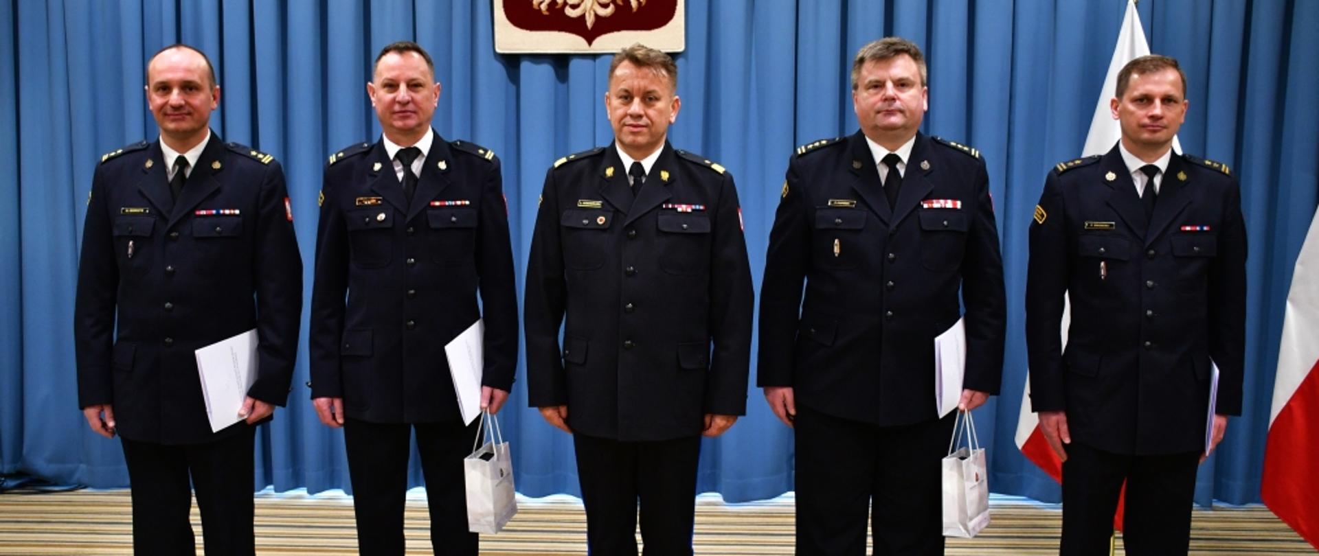 Na zdjęciu widocznych pięciu oficerów PSP w ubraniach wyjściowych. Po środku nadbryg. Jarosław Nowosielski - mazowiecki komendant wojewódzki PSP. Po jego prawej i lewej stronie po dwóch oficerów, trzymających białe teczki z aktami powołania. Po prawej stronie widoczne polskie flagi na stojakach.