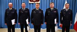Na zdjęciu widocznych pięciu oficerów PSP w ubraniach wyjściowych. Po środku nadbryg. Jarosław Nowosielski - mazowiecki komendant wojewódzki PSP. Po jego prawej i lewej stronie po dwóch oficerów, trzymających białe teczki z aktami powołania. Po prawej stronie widoczne polskie flagi na stojakach.