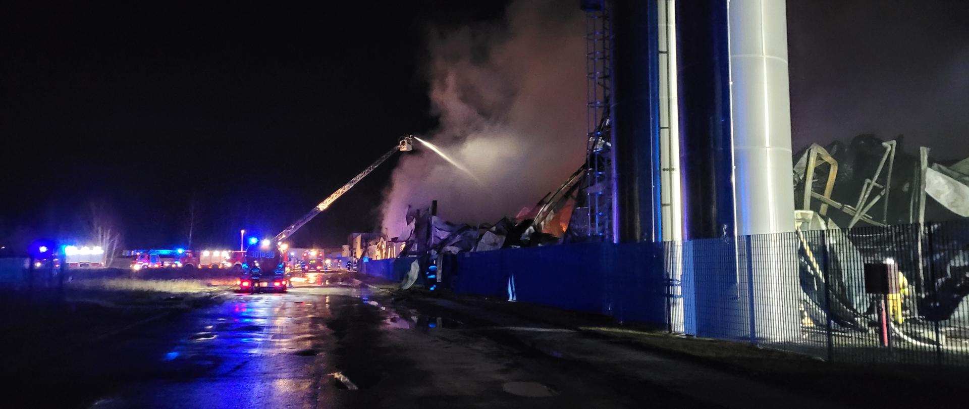 Podawanie wody na dymiący się dach hali produkcyjnej z mechanicznej drabiny pożarniczej, pora nocna, w tle samochody pożarnicze z włączonymi niebieskimi sygnałami świetlnymi 