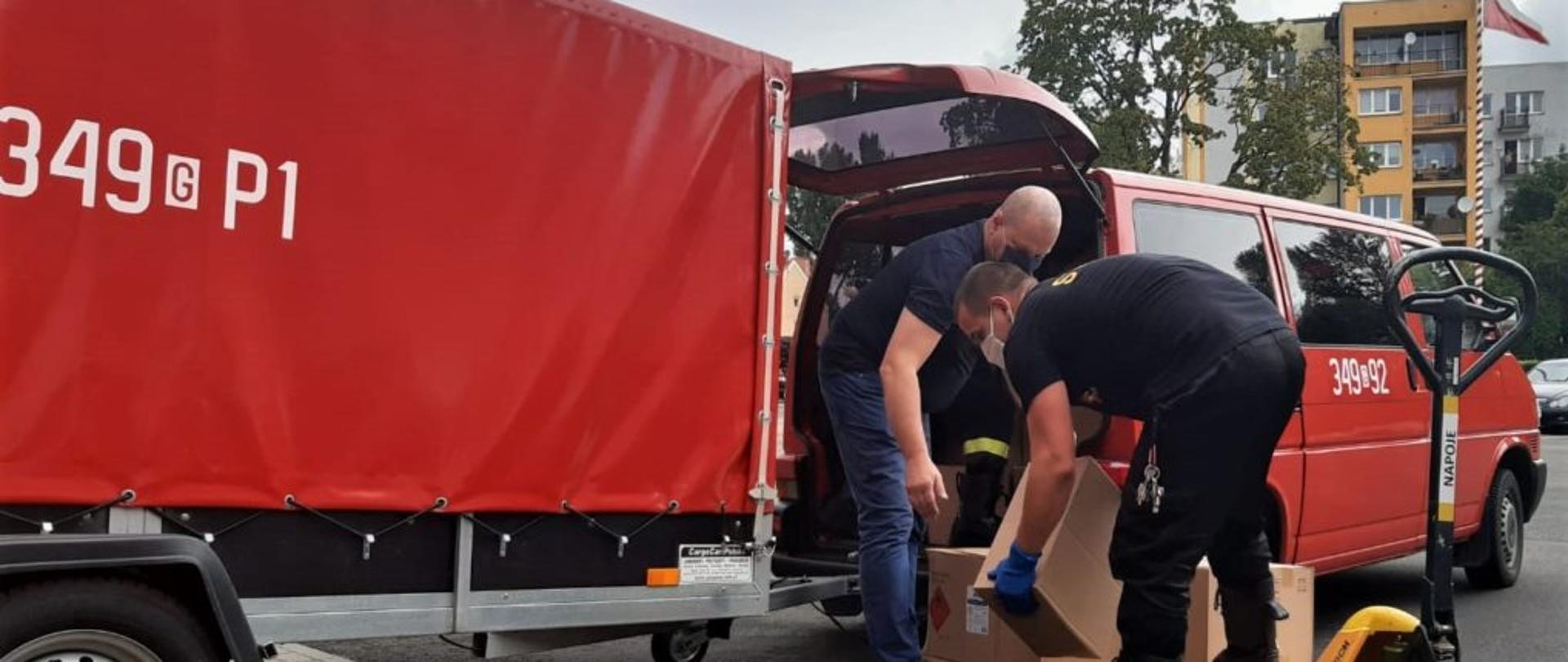 Zdjęcie przedstawia funkcjonariuszy PSP przeładowujących środki ochronne z palety znajdującej się na wózku transportowym do przybyłego samochodu OSP. 