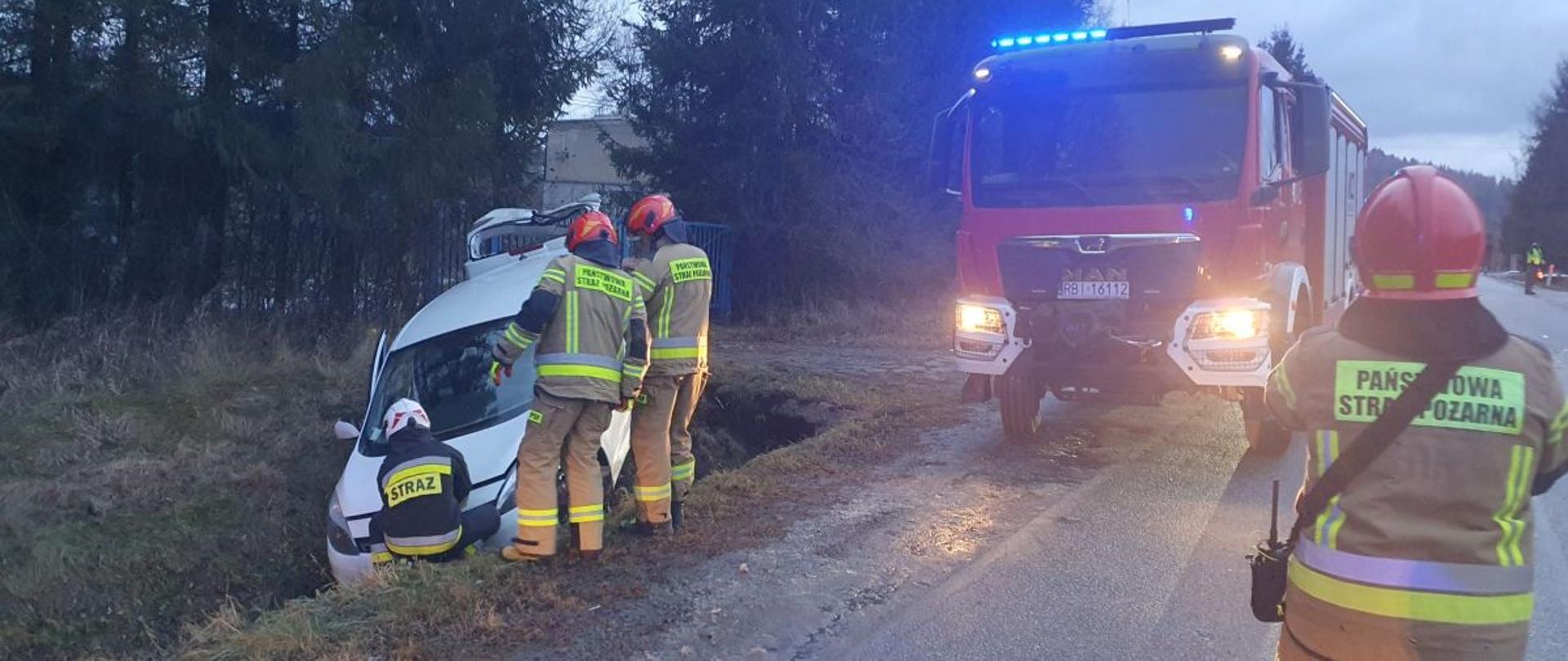 Zdjęcie zawiera samochód w rowie przy którym znajdują się strażacy oraz wóz strażacki zaparkowany na poboczu a także idącego w jego kierunku strażaka.