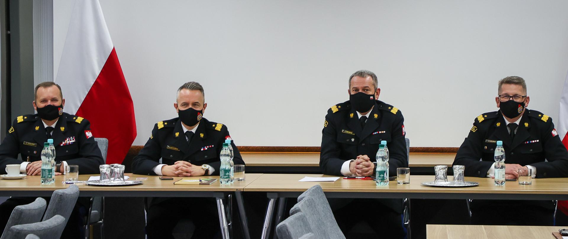 Na zdjęciu widać czterech strażaków, którzy siedzą za stołem prezydialnym. W tle stoją dwie flagi Polski.