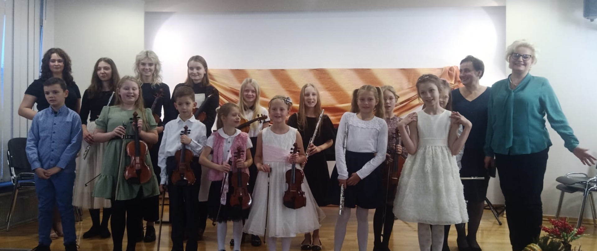 Zdjęcie przedstawia uczniów odświętnie ubranych, którzy trzymają swoje instrumenty skrzypce i flety. Dzieci stoją w dwóch rzędach. Po prawej stronie dwie kobiety. Jedna jest blondynką, a druga brunetką. Blondynka ma zieloną bluzkę, czarne spodnie i okulary. Natomiast brunetka ciemną bluzkę i czarną spódnicę.