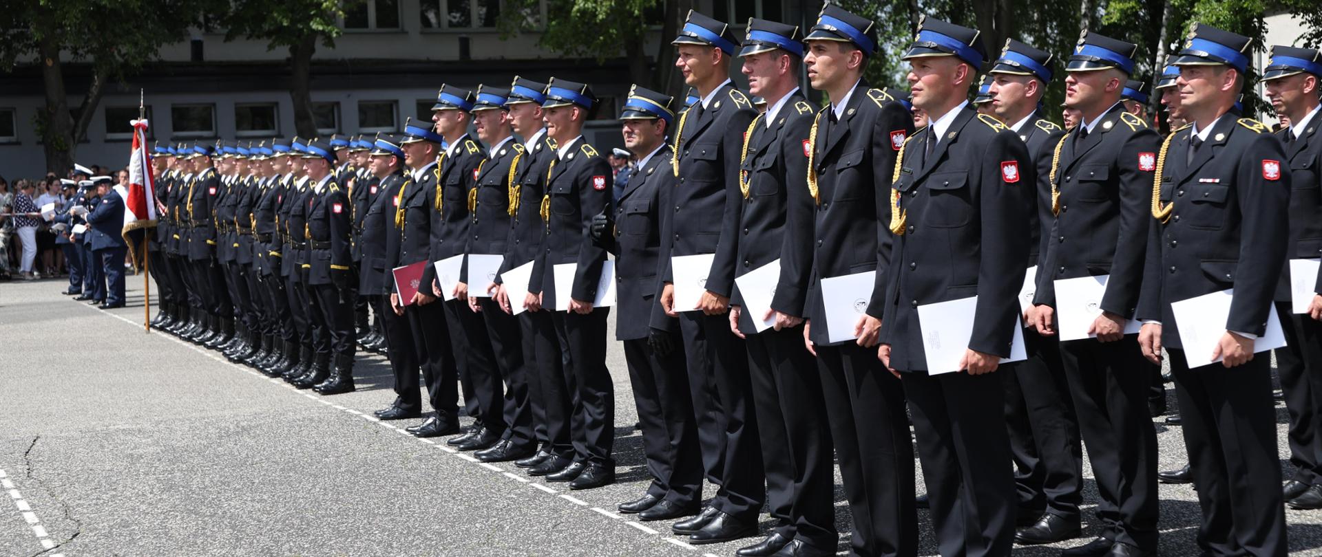 W głębi od lewej stojący poczet sztandarowy następnie promowane kompanie kadetów XXVI turnusu Dziennego Studium Aspirantów CS PSP oraz absolwentów Kwalifikacyjnego Kursu Zawodowego