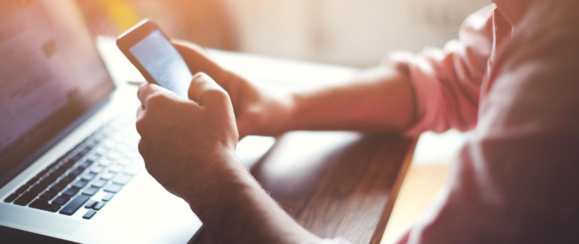 Męskie dłonie dotykają smartfona a obok otwarty laptop leży na biurku