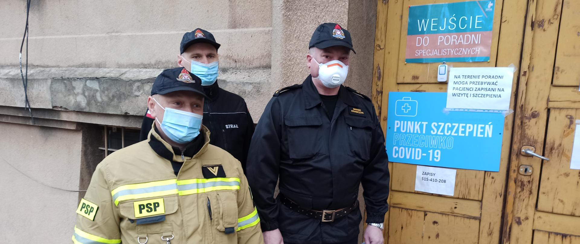 Trzech strażaków w mundurach stoi przed punktem szczepień. Każdy z nich ma na twarzy maseczkę ochronną.