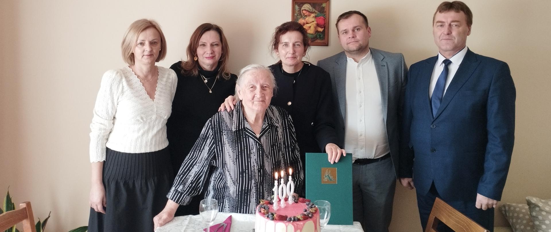 Starsza Pani stoi w pokoju przy stole, na którym stoi tort z palącymi się świeczkami w kształcie liczby 100. Za nią stoją trzy kobiety po lewej i dwóch mężczyzn po prawej.