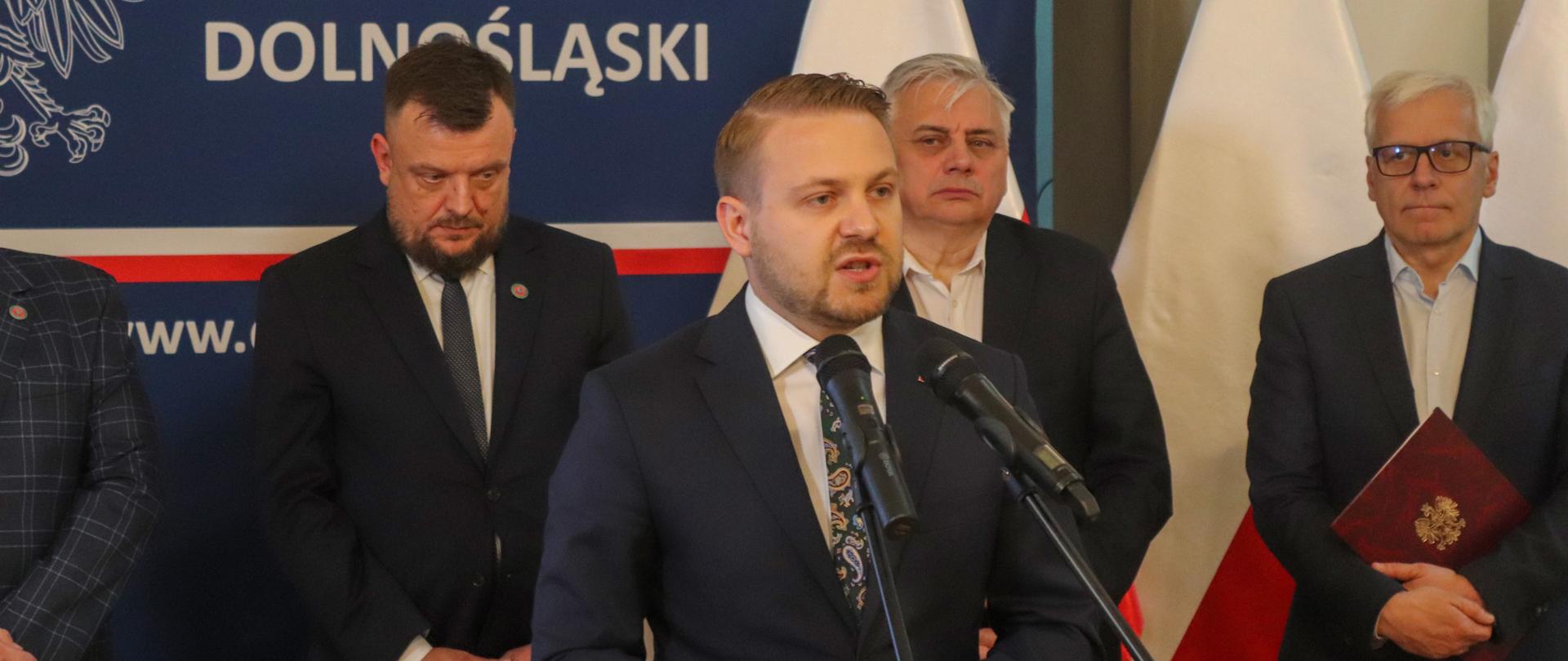 Grupa mężczyzn stojąca na tle flag Polskich 