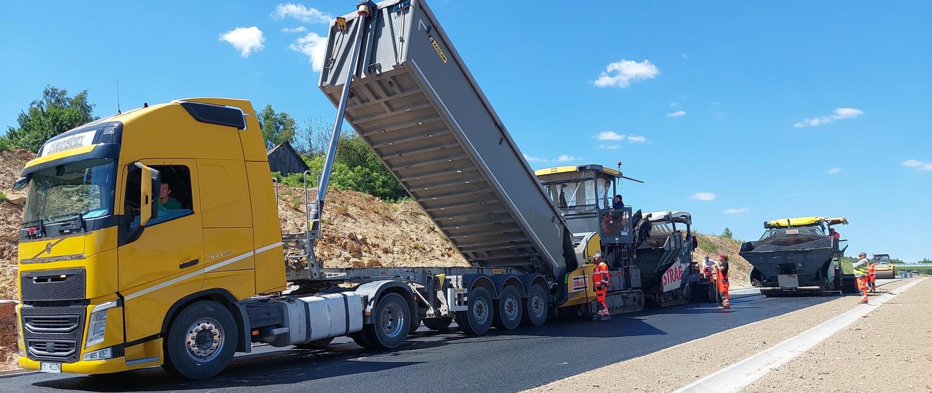 Budowa obwodnicy Wąchocka - układanie nawierzchni, czarna droga asfaltowa, żółty samochód ciężarowy z podniesioną naczepą, za nim maszyny drogowe - układarki,. walec. Pracownicy w pomarańczowych ubraniach ochronnych 