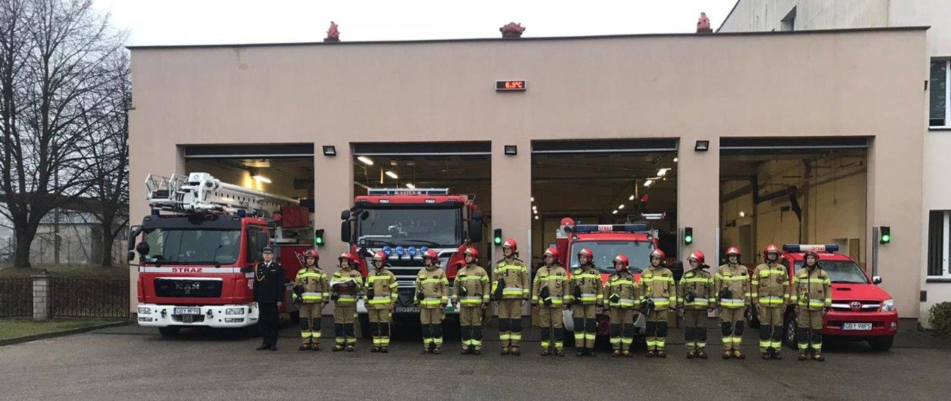 Na zdjęciu widać strażaków ustawionych w szyku na tle samochodów pożarniczych i bram wjazdowych do garażu. Strażacy ubrani w ubrania specjalne koloru piaskowego w hełmach koloru czerwonego.