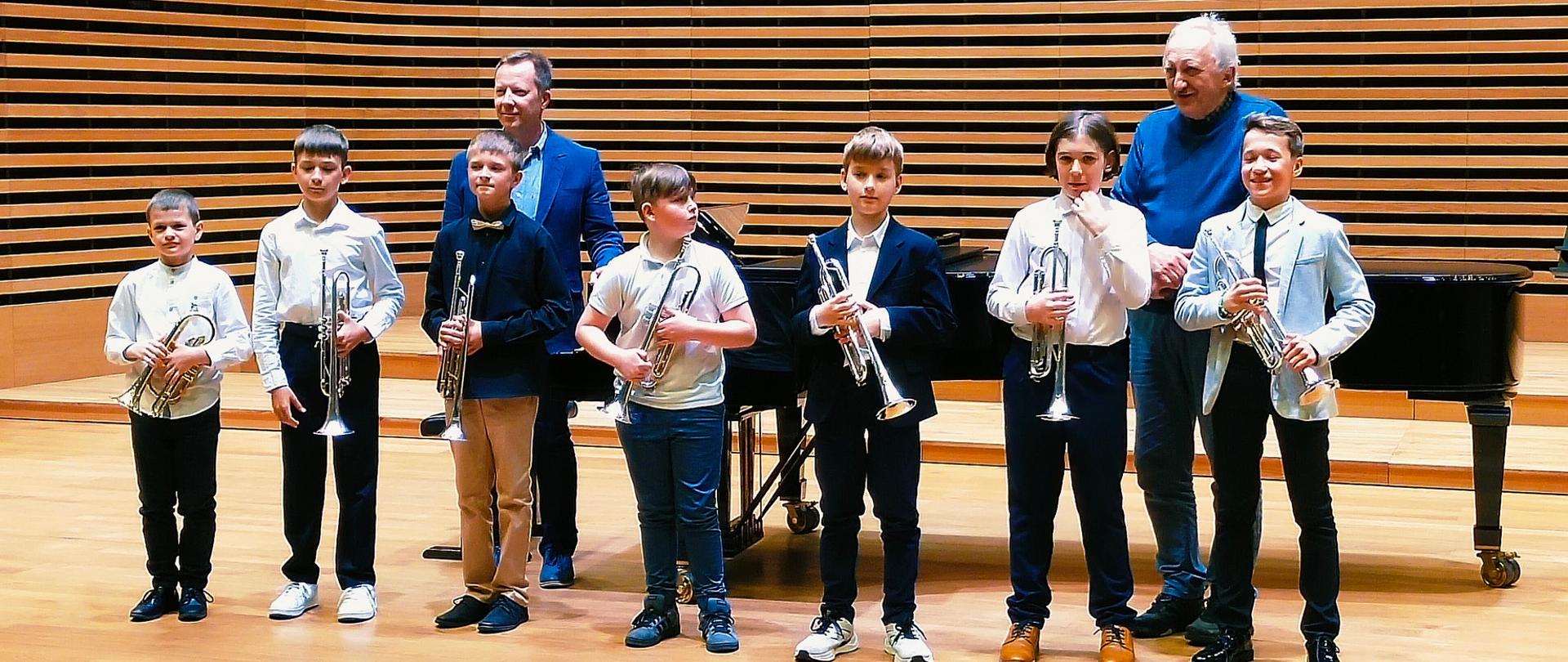 Siedmiu chłopców stoi na scenie z trąbkami, za nimi stoi dwóch mężczyzn.