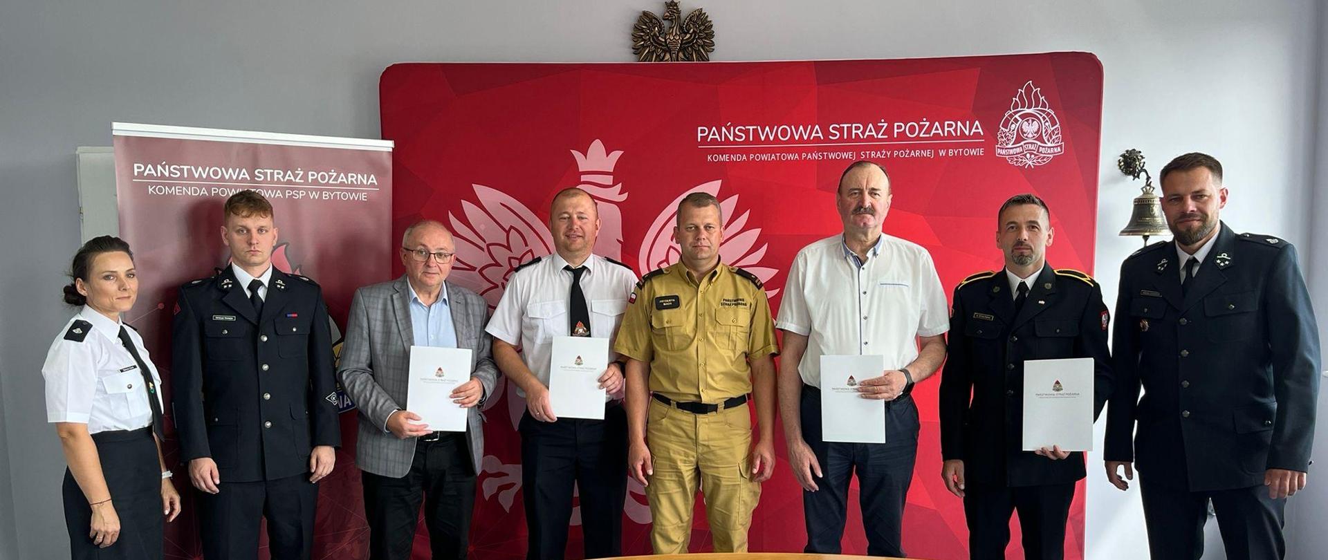 Pięciu mężczyzn w mundurach, dwóch w garniturach i jedna kobieta w mundurze OSP pozują do zdjęcia na tle czerwonego baneru Państwowa Straż Pożarna 