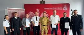 Pięciu mężczyzn w mundurach, dwóch w garniturach i jedna kobieta w mundurze OSP pozują do zdjęcia na tle czerwonego baneru Państwowa Straż Pożarna 