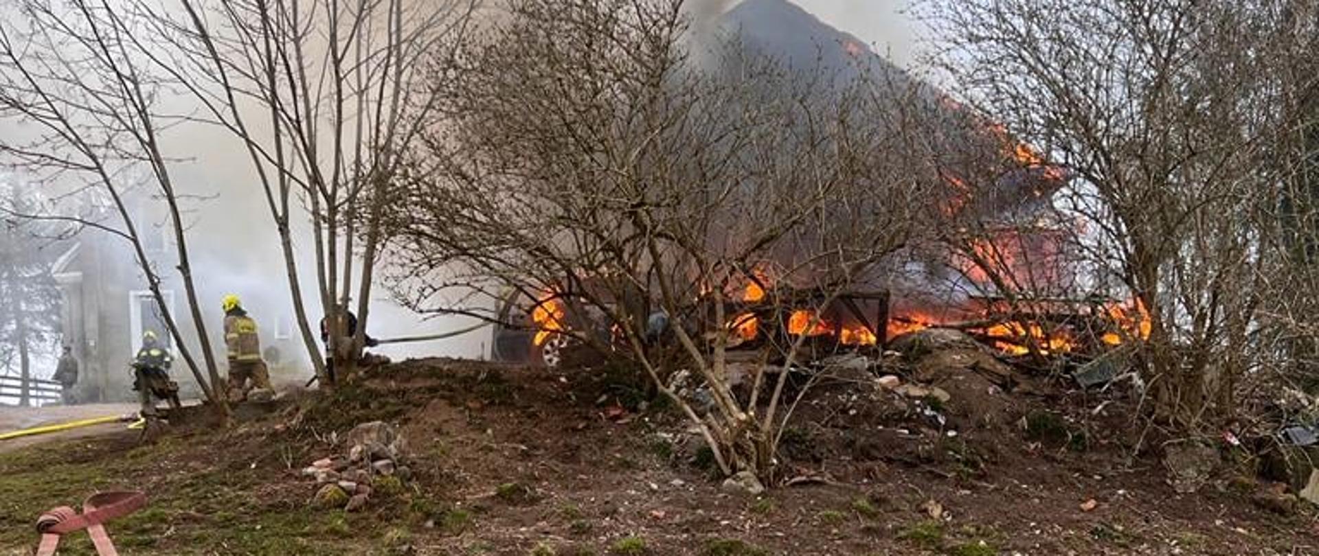 na górce za krzakami widać płomienie palącego się budynku