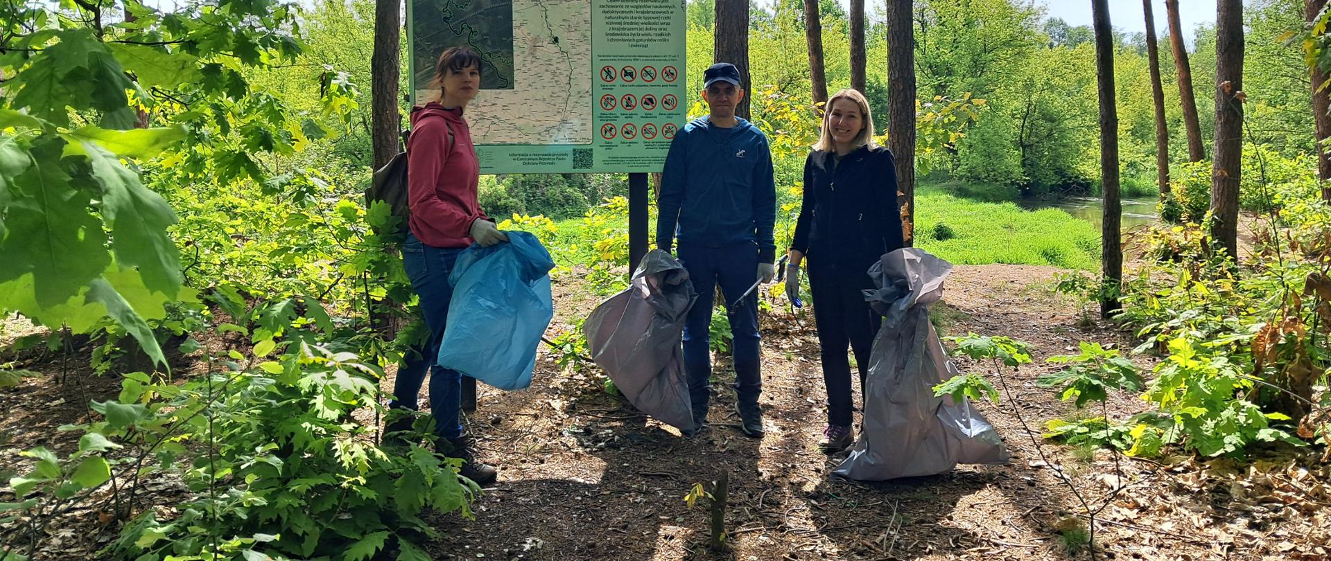 Trójka ludzi w strojach terenowych, z workami na odpady stoi w lesie. Patrzą w obiektyw. Za nimi stoi tablica z napisem Rezerwat Przyrody Rawka, poniżej tablicy, druga tablica z mapą i napisanym tekstem. 