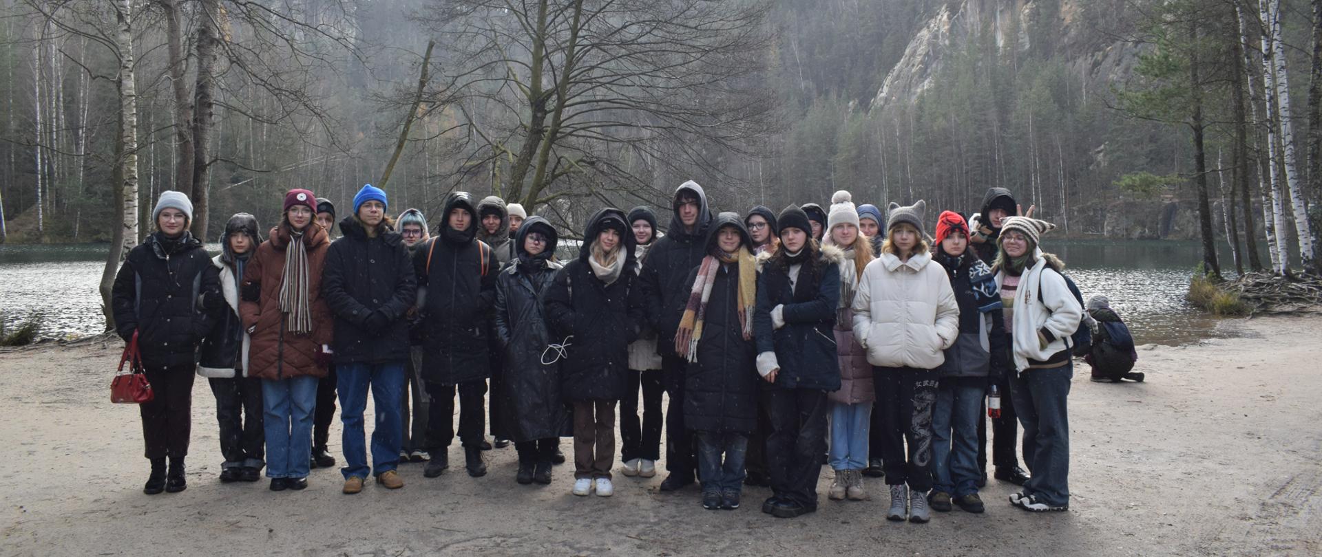Na zdjęciu grupa młodzieży stoi na tle gór, drzew i zbiornika wodnego. Jest listopad. Ludzie są ciepło ubrani.