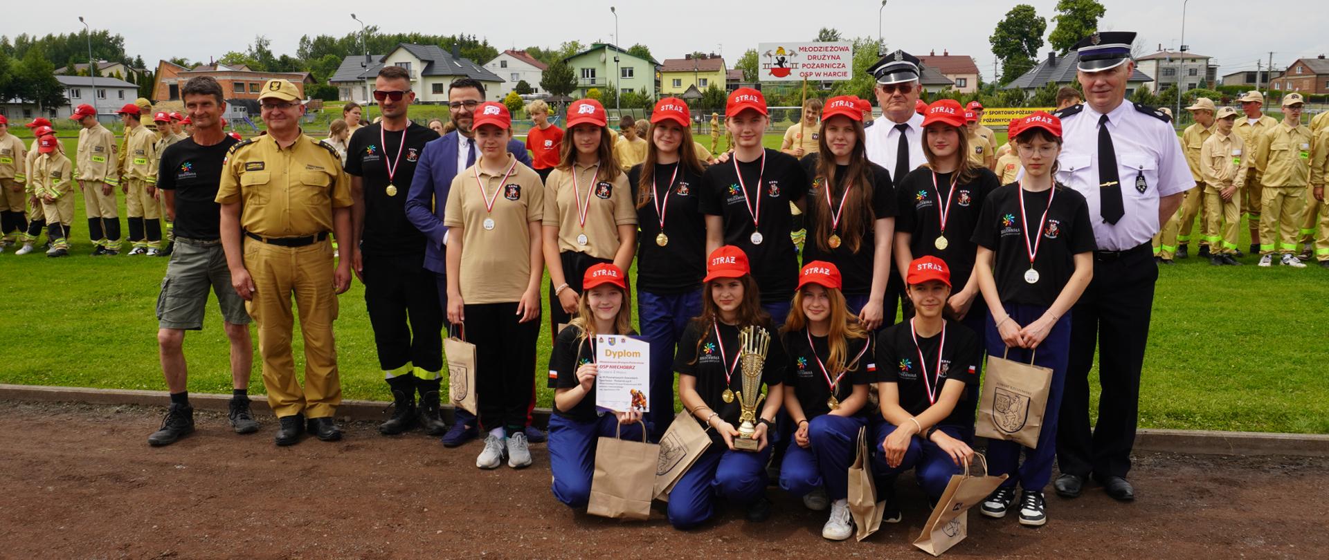 Grupowe zdjęcie młodzieżowej drużyny pożarniczej z opiekunami i przedstawicielami służb. Na pierwszym planie klęczy młodzież w czerwonych czapkach z napisem ‘STRAŻ’, trzymająca dyplom, puchar i torby z nagrodami. W tle widoczni inni uczestnicy zawodów ubrani w mundury oraz transparent z napisem ‘Młodzieżowa Drużyna Pożarnicza OSP w Harcie