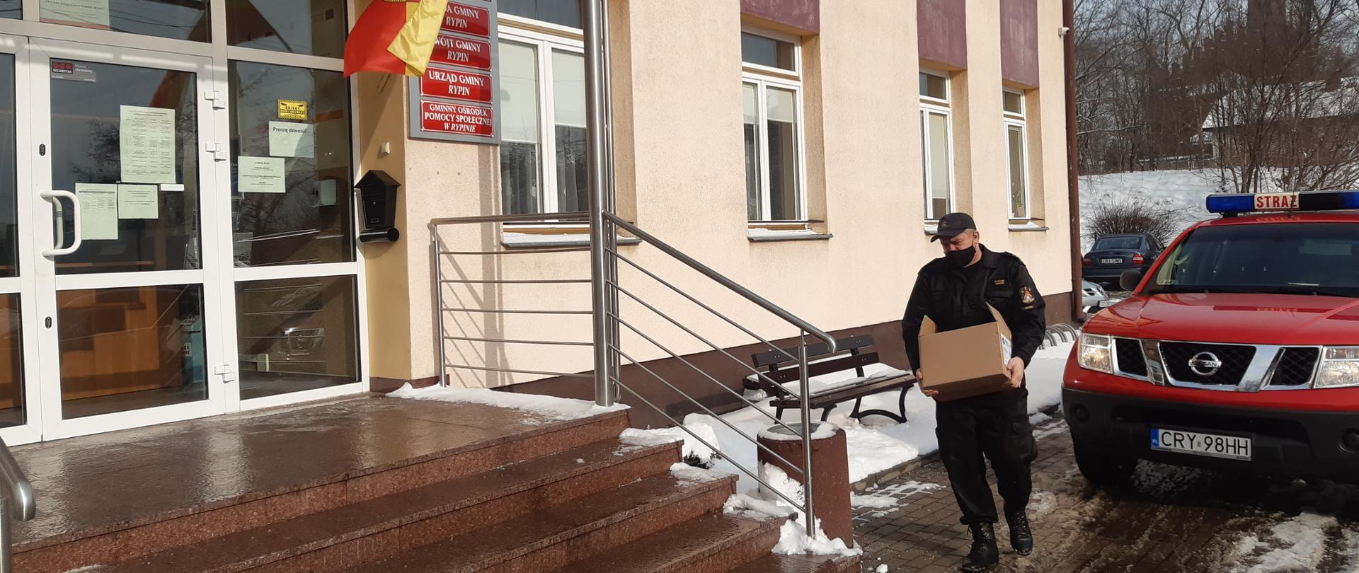 Strażak niesie karton z ulotkami. Przed nim brązowe schodowy po których zaraz wejdzie. Za schodami przeszklone drzwi do budynku. W tle czerwony samochód strażacki. Gdzieniegdzie widoczny śnieg.