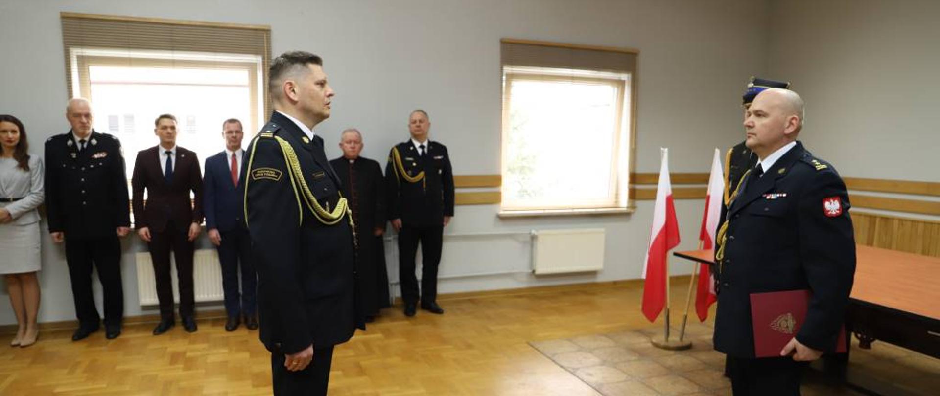 Zdjęcie przedstawia Zastępcę Podlaskiego Komendanta Wojewódzkiego Państwowej Straży Pożarnej brygadiera Marcina Bielakowicza podczas przyjmowania meldunku od nowo powołanego Komendanta Powiatowego Państwowej Straży Pożarnej w Zambrowie młodszego brygadiera Andrzeja Kalinowskiego. Komendanci ubrani w mundury wyjściowe ze sznurem galowym. Uroczystość odbywała się w jednym z pomieszczeń Komendy Powiatowej Państwowej Straży Pożarnej w Zambrowie.