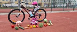na boisku szkolnym stoi rower, rolki i okulary pływackie, na płycie boiska leżą warzywa i owoce
