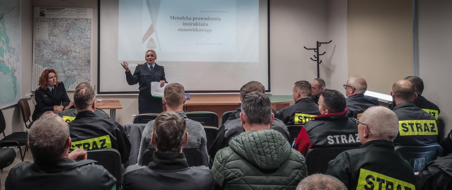 Zdjęcie zrobione w pomieszczeniu. Na pierwszym planie grupa siedzących strażaków ratowników OSP zwróceni plecami, w oddali dwie osoby prowadzące szkolenie, jedna siedzi druga stoi na tle prezentacji multimedialnej z napisem: „Metodyka prowadzenia instruktarzu stanowiskowego”.