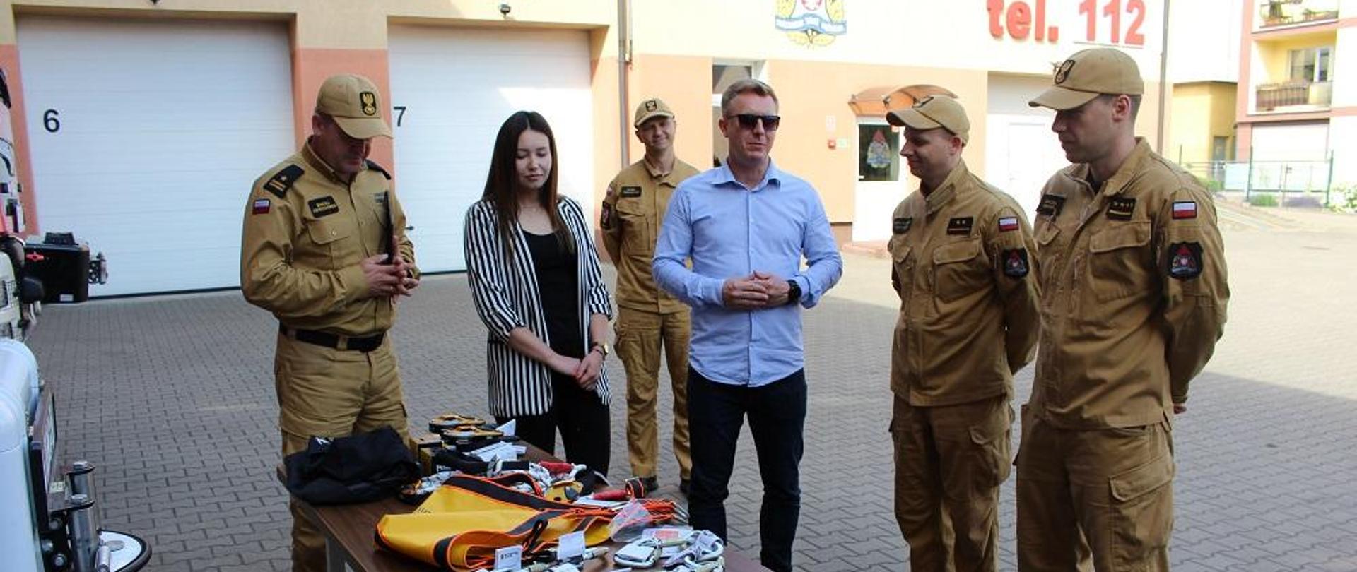 Zdjęcie przedstawia strażaków podczas przekazywania sprzętu ratowniczego.
W tle budynek straży.
