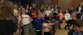 Na dużej sali odbywa się zabawa uczniowska. Dzieci i młodzież przechodzą pod przeciągniętą wszerz sali czerwoną liną. Śmieją się.