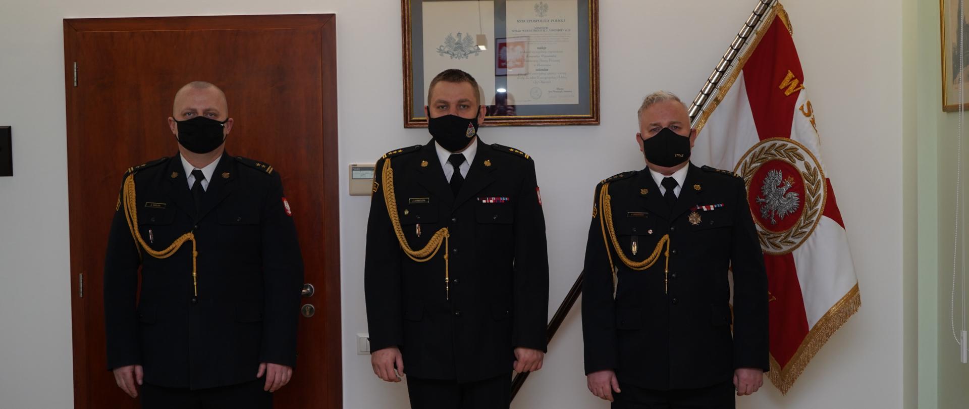 Na zdjęciu widnieją strażacy - komendanci Państwowej Straży Pożarnej w trakcie wręczania aktów nominowania i odwołania ze stanowiska Komendanta Powiatowego PSP w Pile. Uroczystość odbywa się wewnątrz pomieszczenia, oświetlenie sztuczne. 