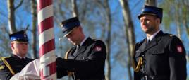 Trzech umundurowanych strażaków stoi przy wysokim maszcie pomalowanym w czerwono-białe pasy. Dwóch z nich obsługuje linę przy maszcie, podnosząc lub przygotowując flagę, trzeci stoi obok na baczność. W tle widoczne są drzewa bez liści i niebieskie niebo, a obok stoi czerwony hydrant. Na dole znajduje się emblemat Państwowej Straży Pożarnej z napisem „Gryfice”.