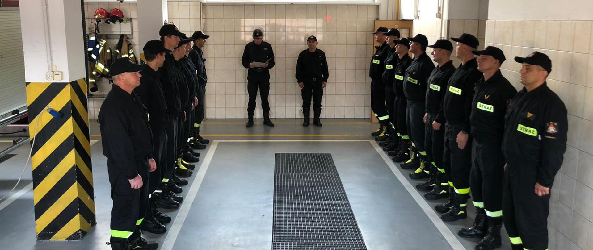 Pomieszczenie garażowe, a w nim strażacy stojący w szeregu po lewej i prawej stronie, w centrum stoi dwóch strażaków, jeden z nich czyta trzymane dokumenty, drugi stoi na baczność